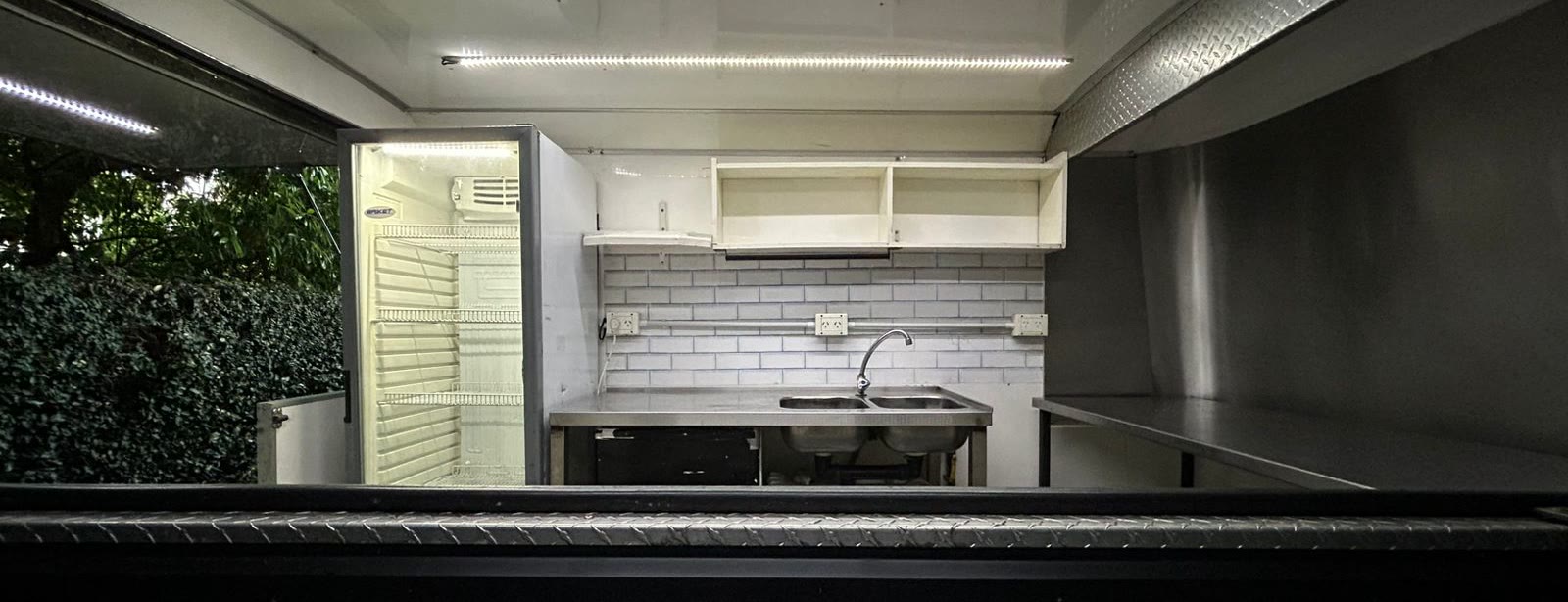 Interior del trailer equipado de noche con las tiras LED prendidas: heladera exhibidora, bacha doble con grifería, mesada de acero y la barra colgante exterior en primer plano
