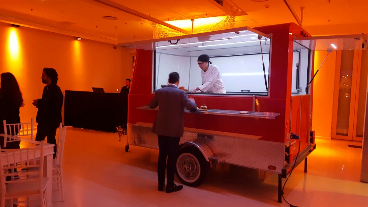 Un cocinero de chaqueta blanca atiende desde la ventana del food truck a un invitado de traje, en un evento corporativo puertas adentro