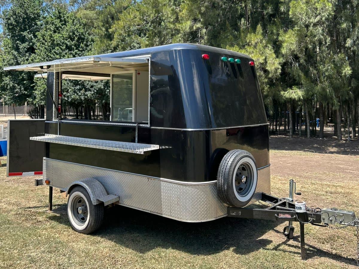 Trailer gastronómico negro de OCHO con el portón abierto hacia arriba, la barra colgante desplegada y la lanza de remolque con rueda jockey a la vista