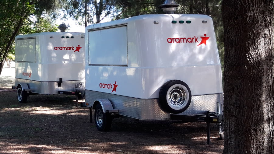 Dos trailers de OCHO ploteados con la gráfica de una marca gastronómica, blancos con el logotipo en rojo, en un pinar
